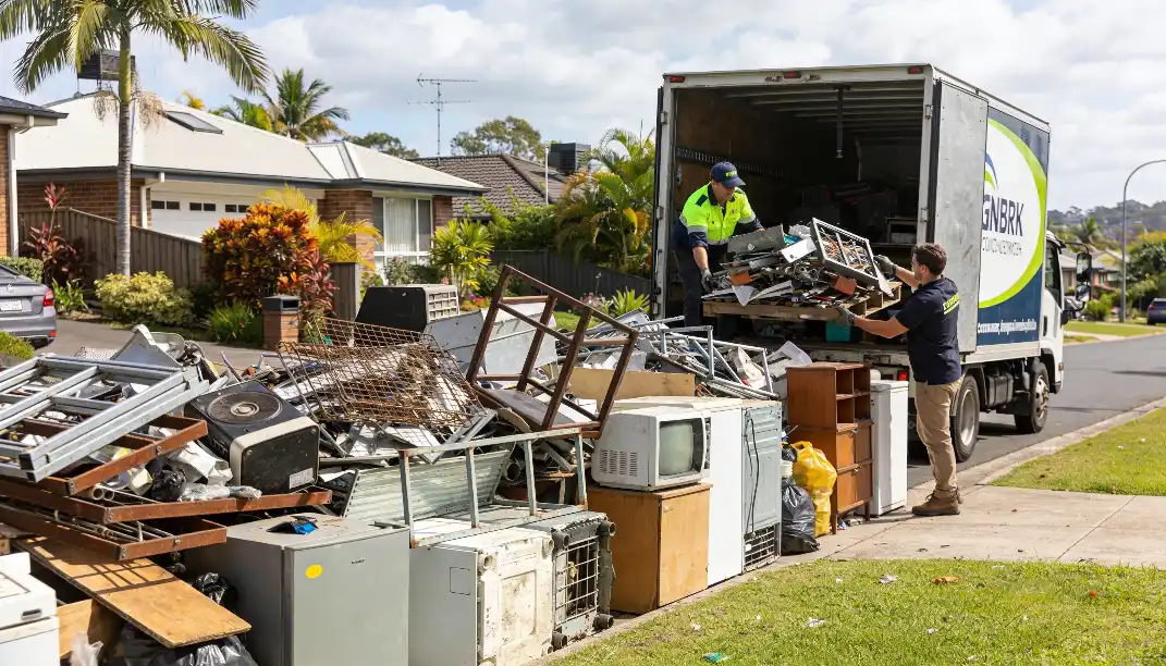 Two workers in hi-vis vests loading large amounts of bulk waste including old televisions furniture and scrap metal into a council collection truck parked on a Brisbane suburban street surrounded by palm trees and residential homes.